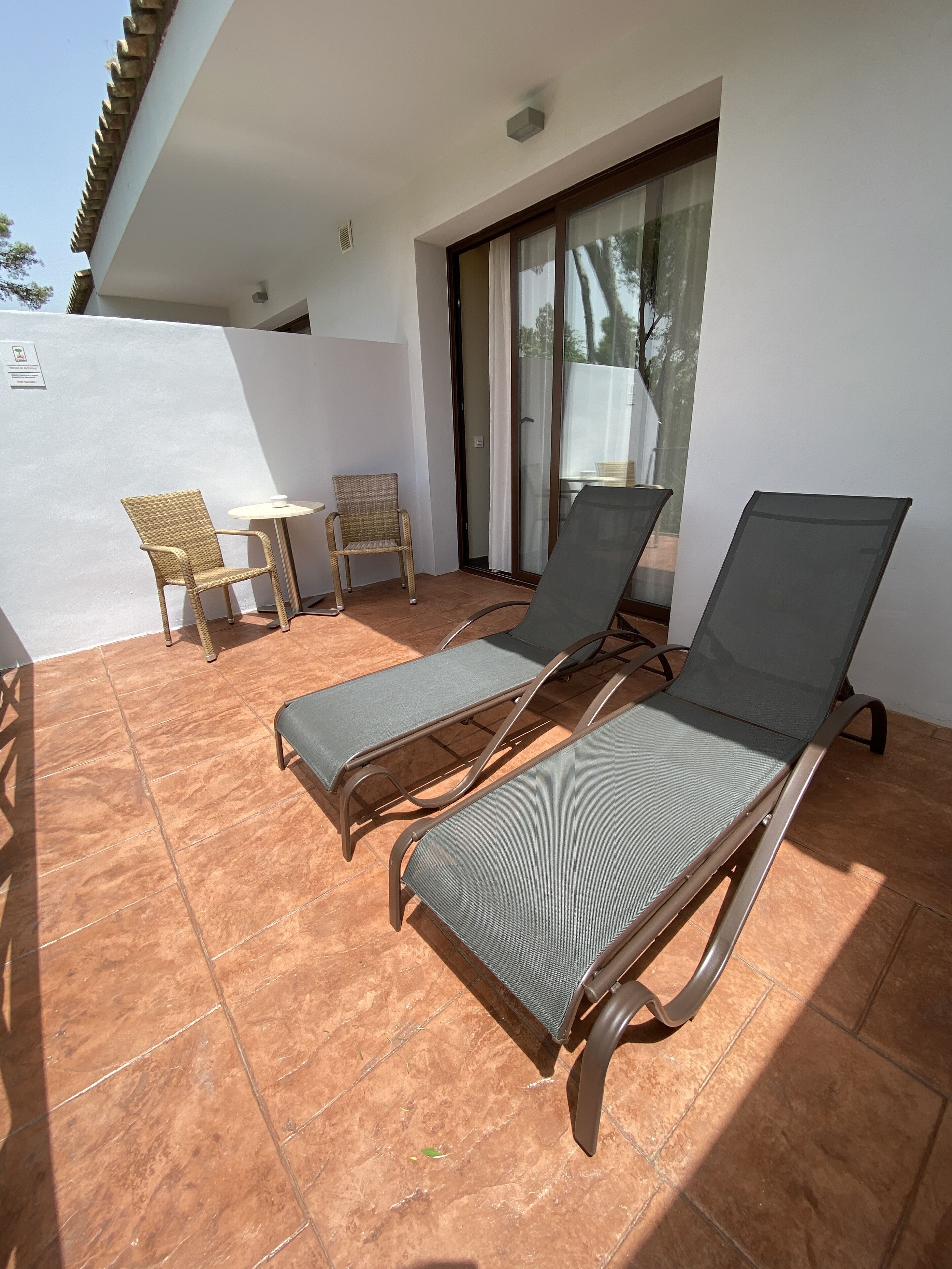 A sunlit outdoor seating area with two lounge chairs and a small table, a wicker chair, and a glass door leading to a covered patio.