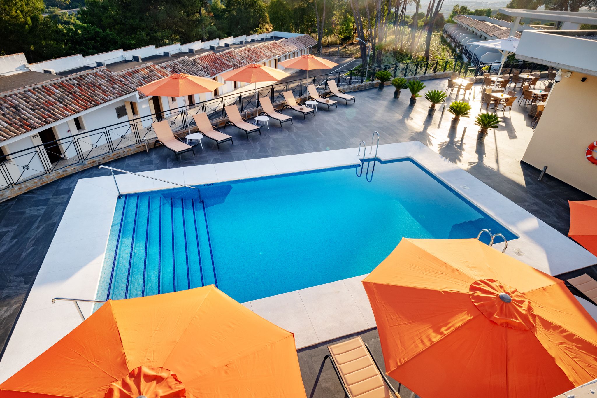A rooftop pool area with lounge chairs and umbrellas, surrounded by a white building with a tiled roof.