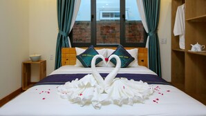 A neatly made bed with a swan-shaped towel arrangement, a wooden headboard, and a view of a brick wall through a window with curtains. - Home Sweet Home Villa (Da Nang)