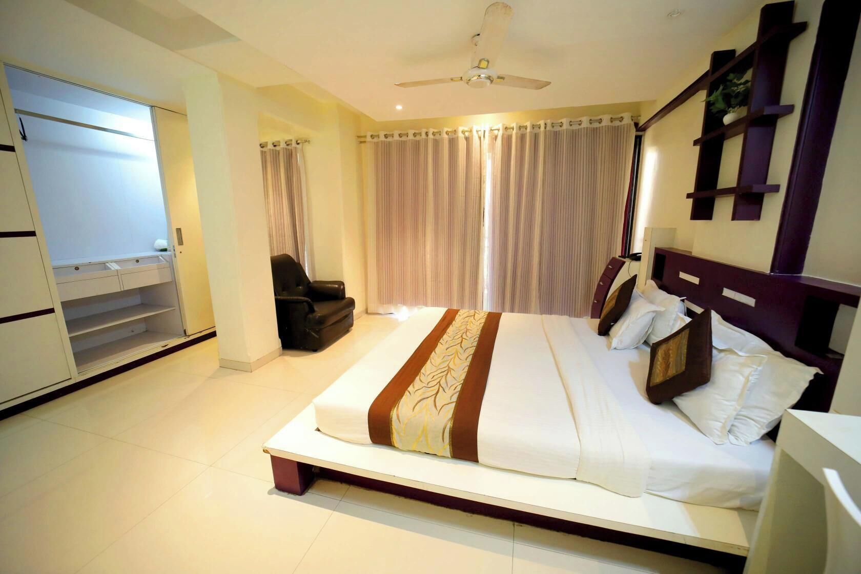 A hotel room with a bed, a chair, a wardrobe, and a ceiling fan.