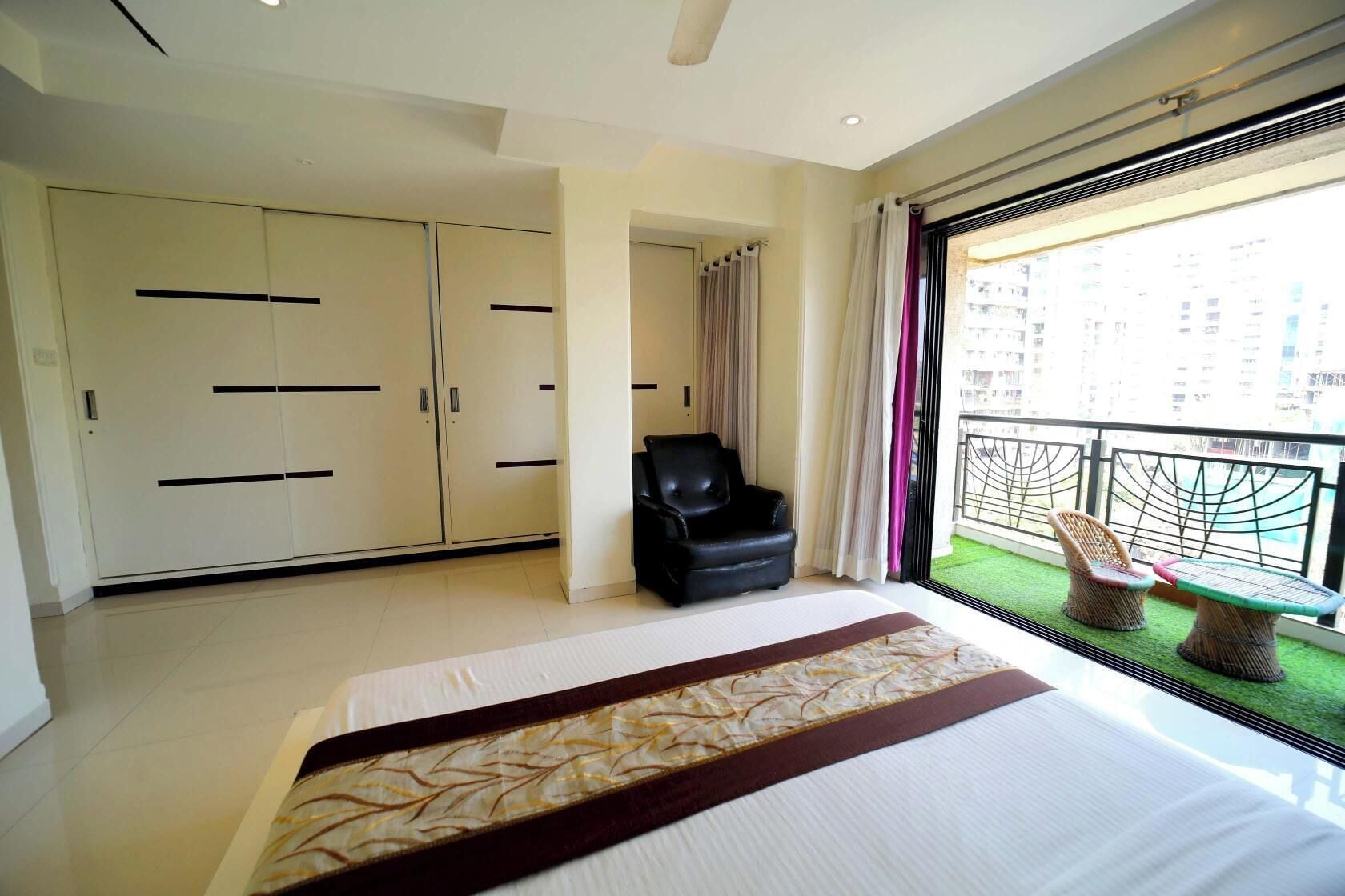 A hotel room with a large bed, a black leather chair, a balcony with wicker furniture, and a view of a cityscape.