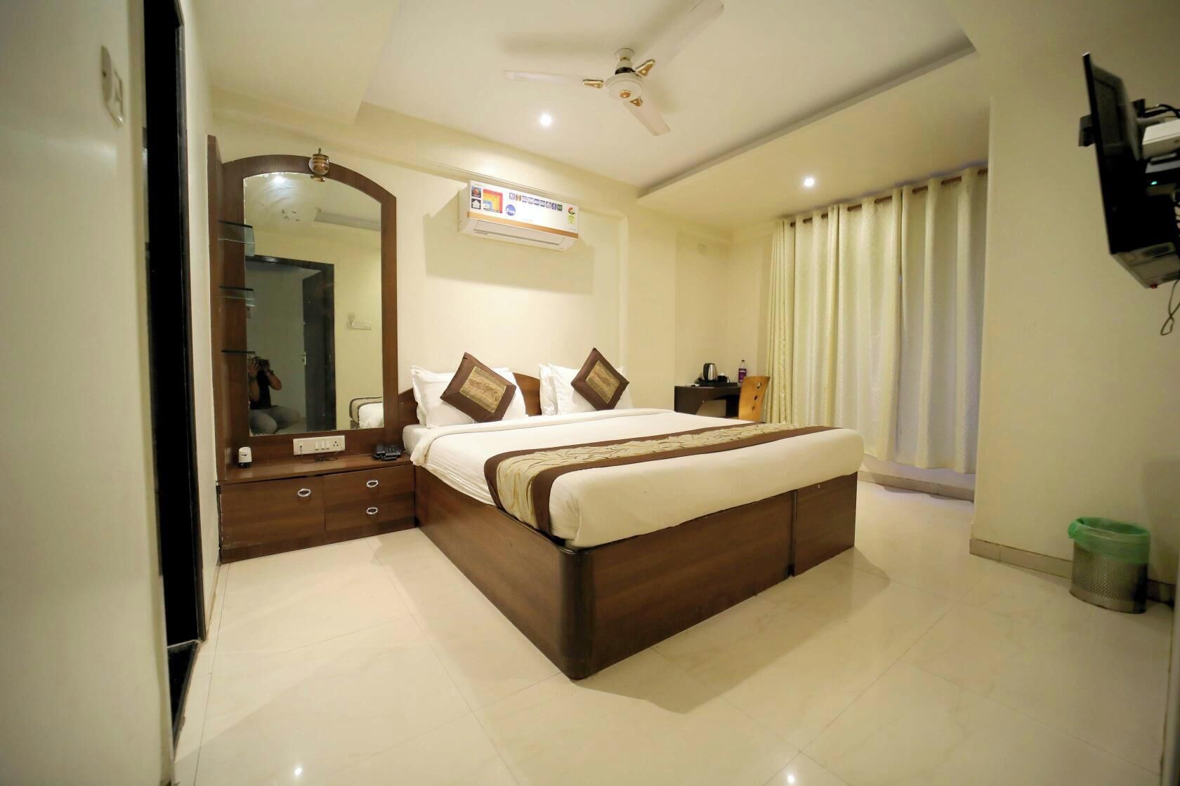 A hotel room with a large bed, a bedside table, a mirror, a ceiling fan, and a window with curtains.