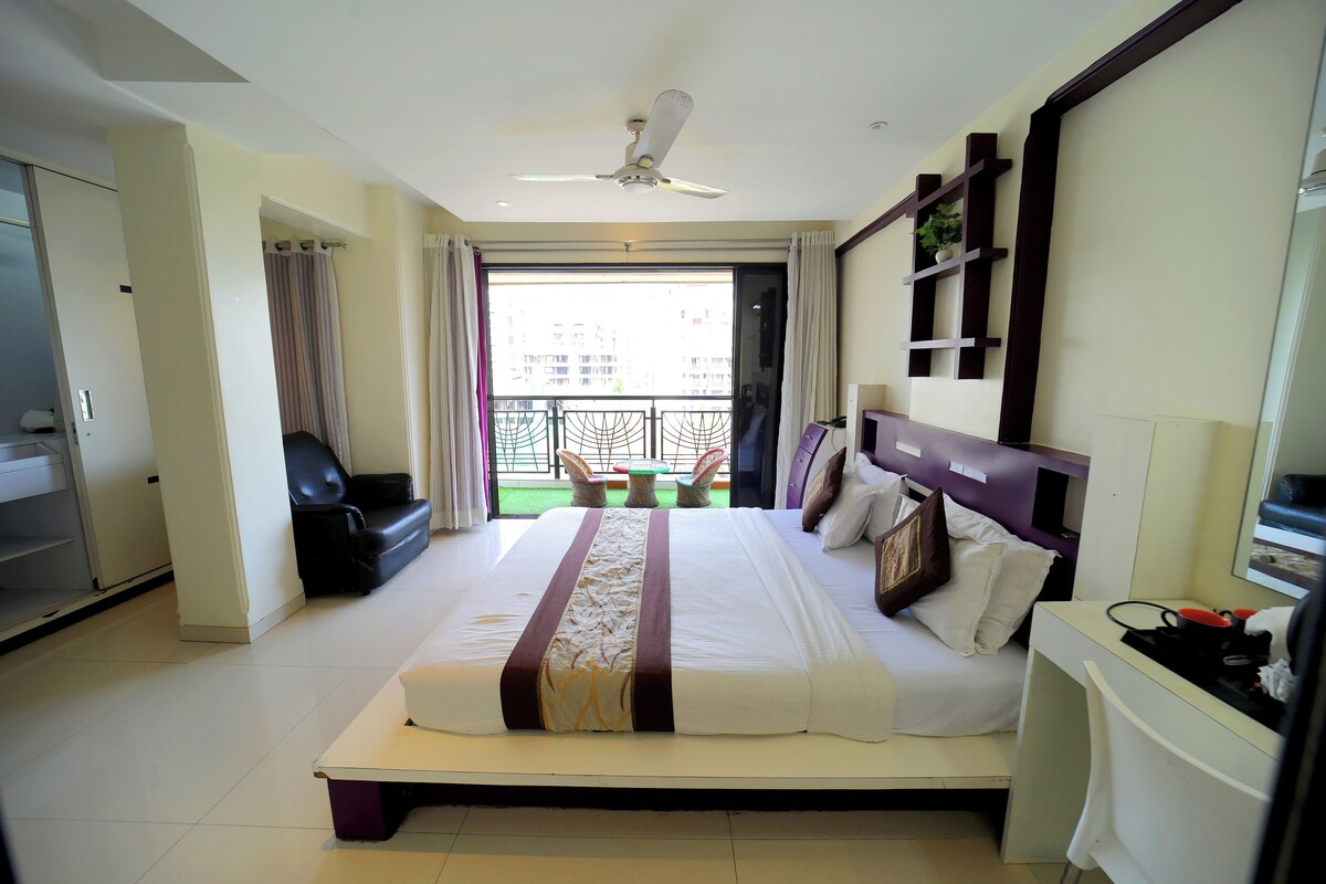 A hotel room with a large bed, a desk, a chair, a ceiling fan, and a balcony with a view of buildings.