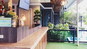 Een receptiegedeelte met een houten balie, een aan de muur bevestigde televisie, een potplant en uitzicht op het buitenbalkon met groen.