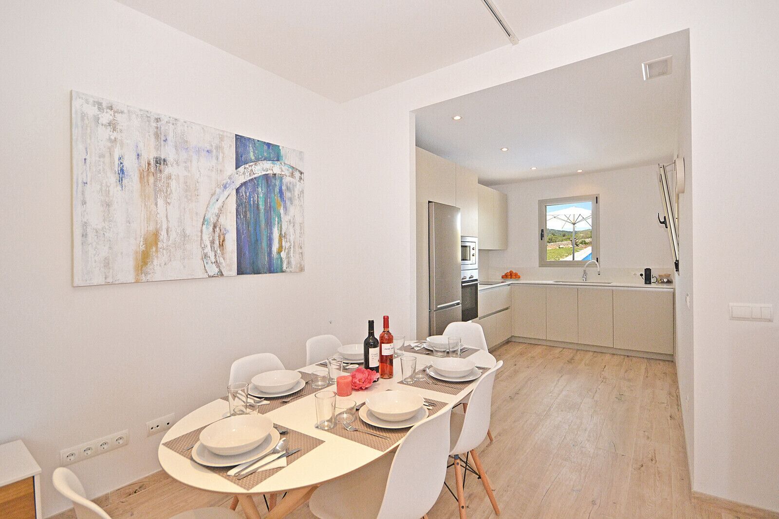 Un comedor moderno con una mesa preparada para cuatro personas, una cocina al fondo y arte abstracto en la pared.