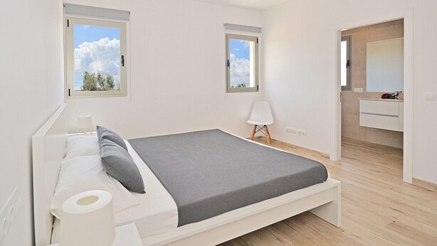 A modern bedroom with a bed, a small bedside table, a chair, and a bathroom visible through an open door.