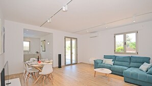 A modern living room with a blue sofa, a dining area with white chairs, and a kitchen visible through an open door.