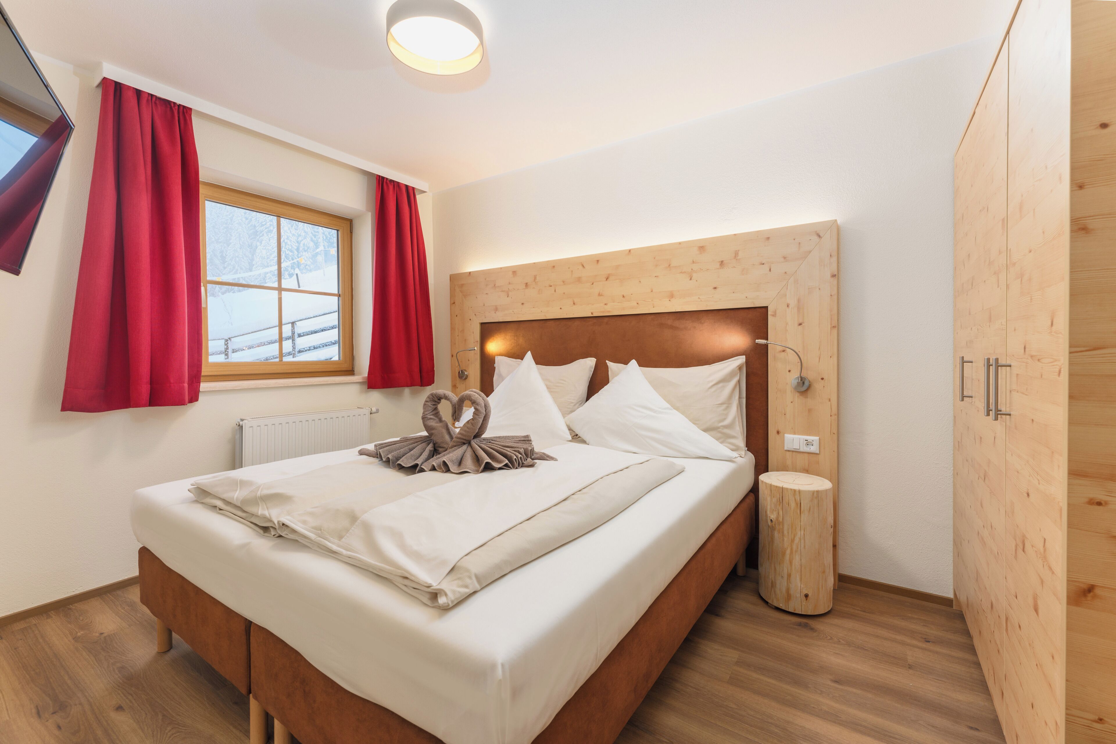 Una cama bien hecha con cabecera de madera, una mesita de noche y una ventana con cortinas rojas.