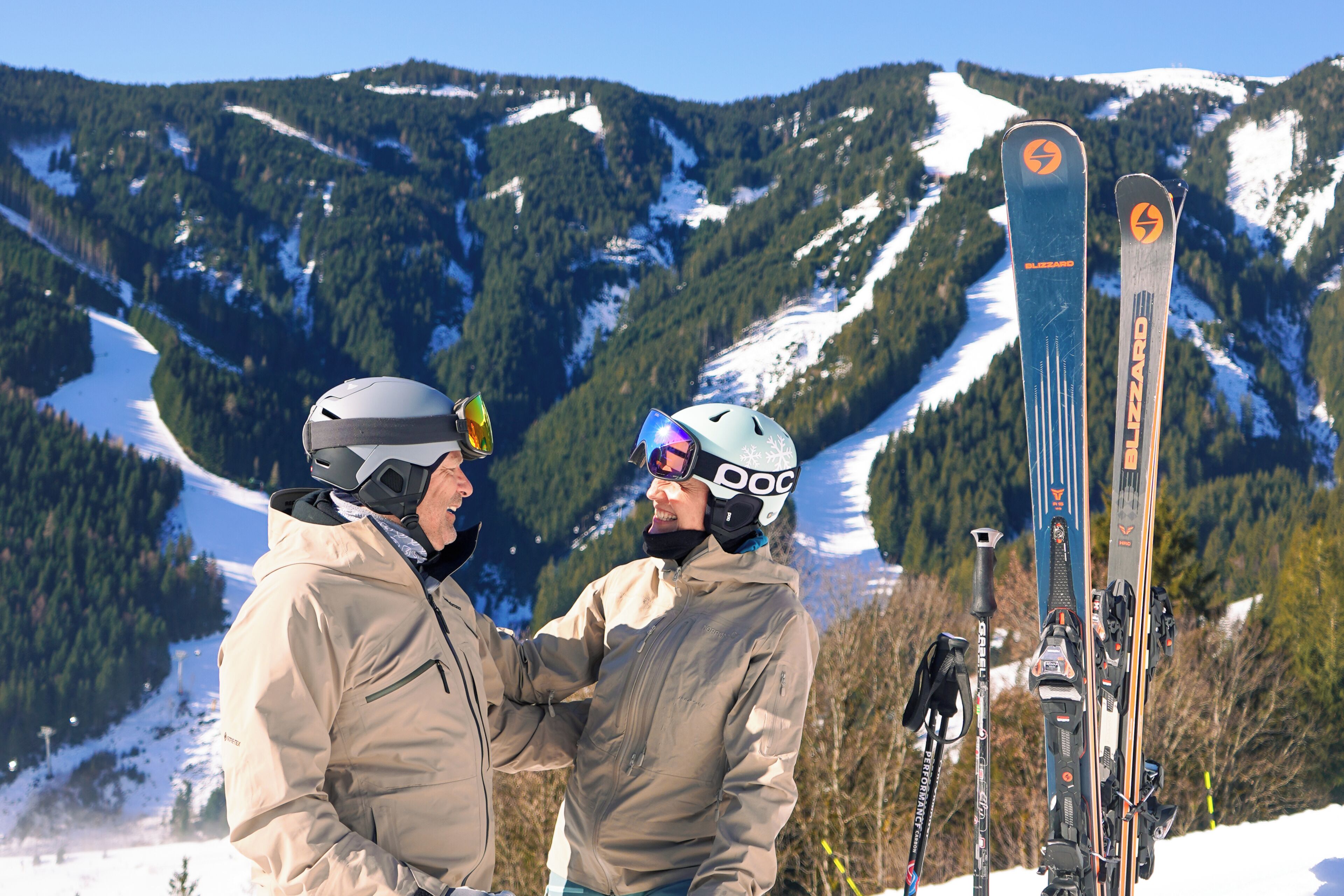 Two people standing with skis and ski poles, with snow-covered mountains in the background.
