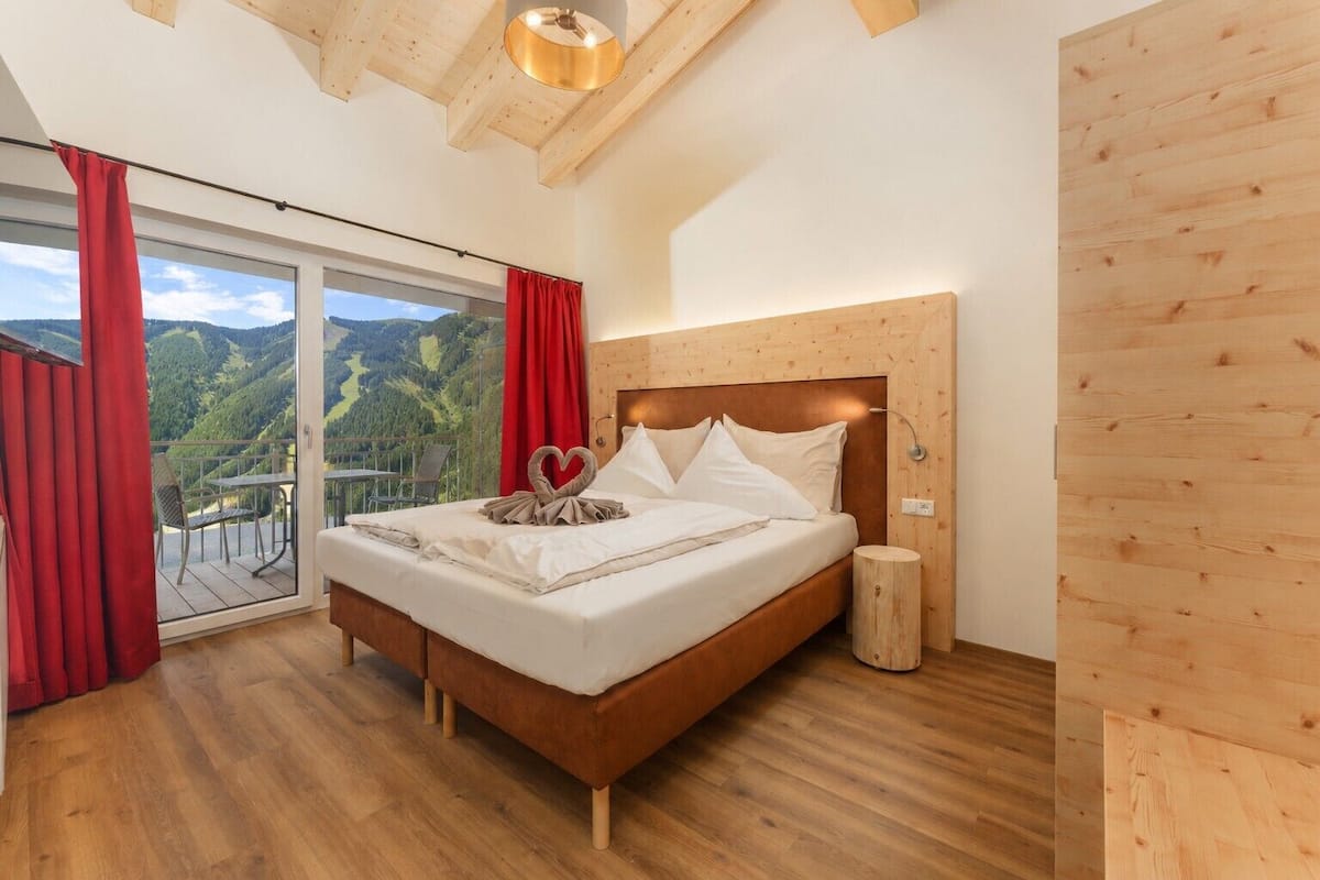 Une chambre à coucher avec une tête de lit en bois, un lit recouvert de draps blancs, une serviette en forme de cœur et une vue sur un paysage montagneux, visible à travers une porte-fenêtre coulissante.