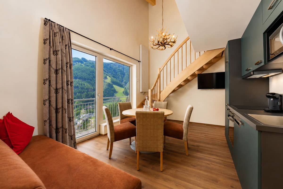 Une cuisine moderne dotée d’un four encastré, un coin repas avec une table et des chaises, un canapé et une grande fenêtre donnant sur un paysage boisé.