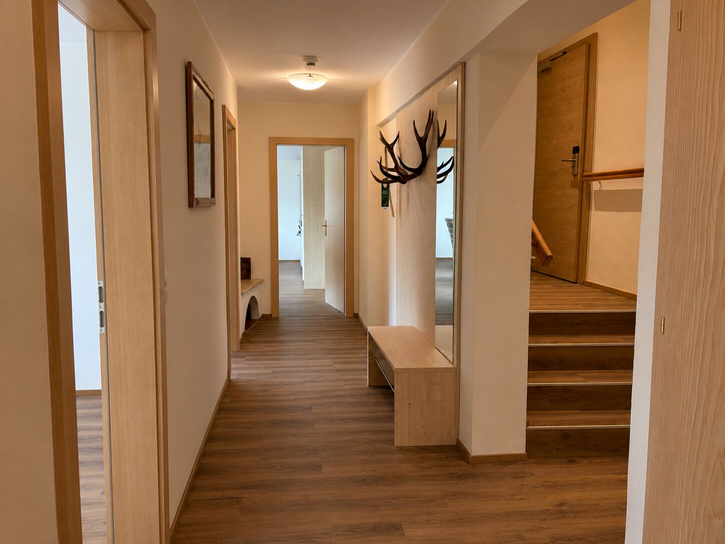 A hallway with wooden flooring, a bench, a staircase, and a door with a mirror.