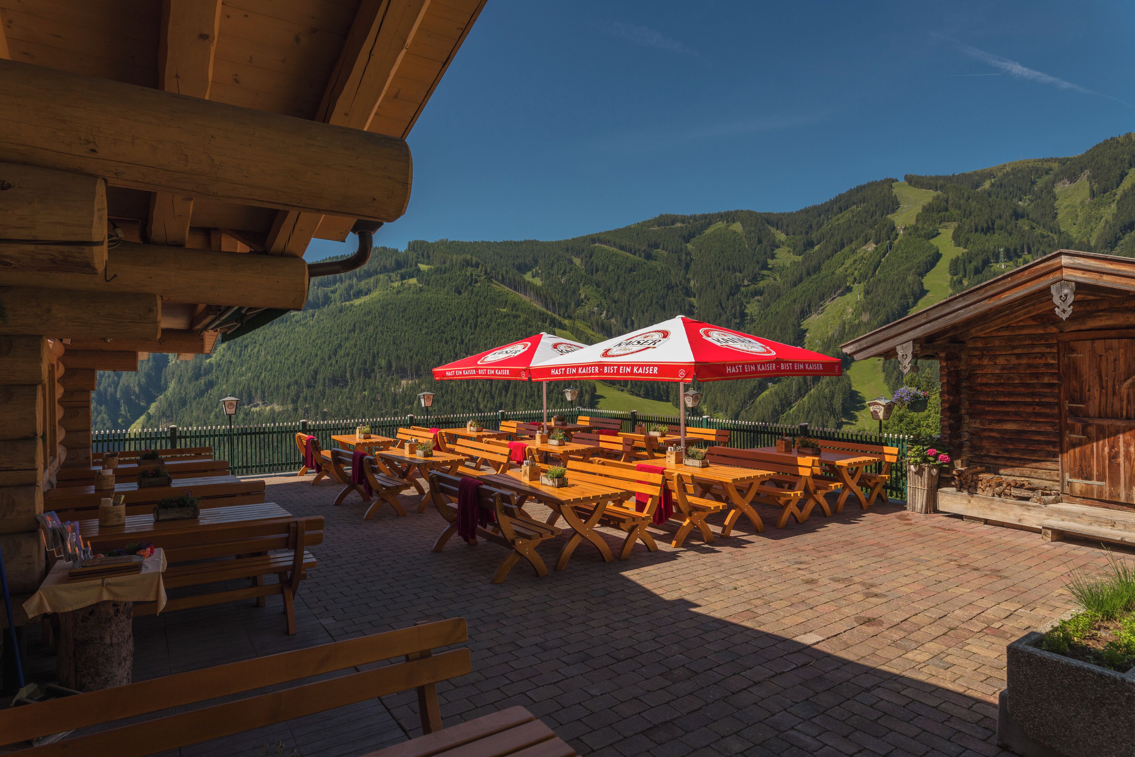 An outdoor seating area with wooden tables and benches, red umbrellas, and a view of green hills.