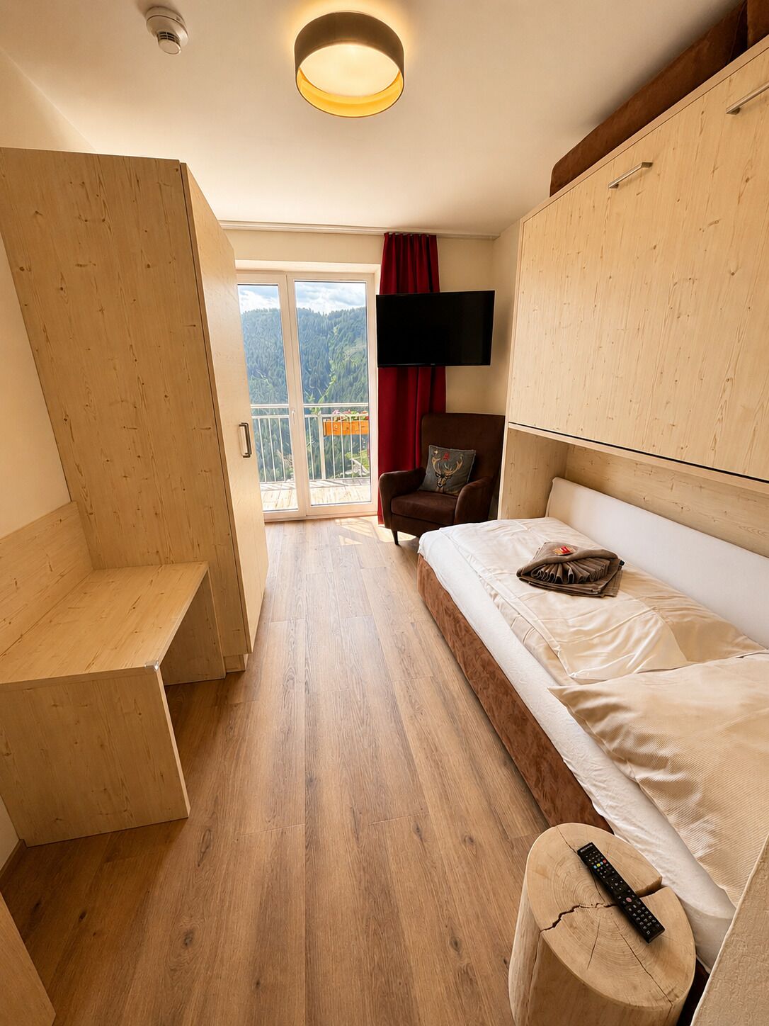 A hotel room with a bed, a TV mounted on the wall, a wooden bench, and a balcony with a view of the mountains.