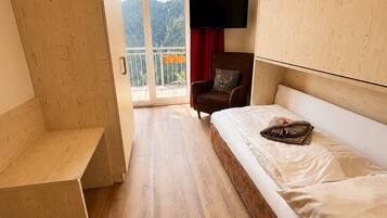 A hotel room with a bed, a TV mounted on the wall, a wooden bench, and a balcony with a view of the mountains.