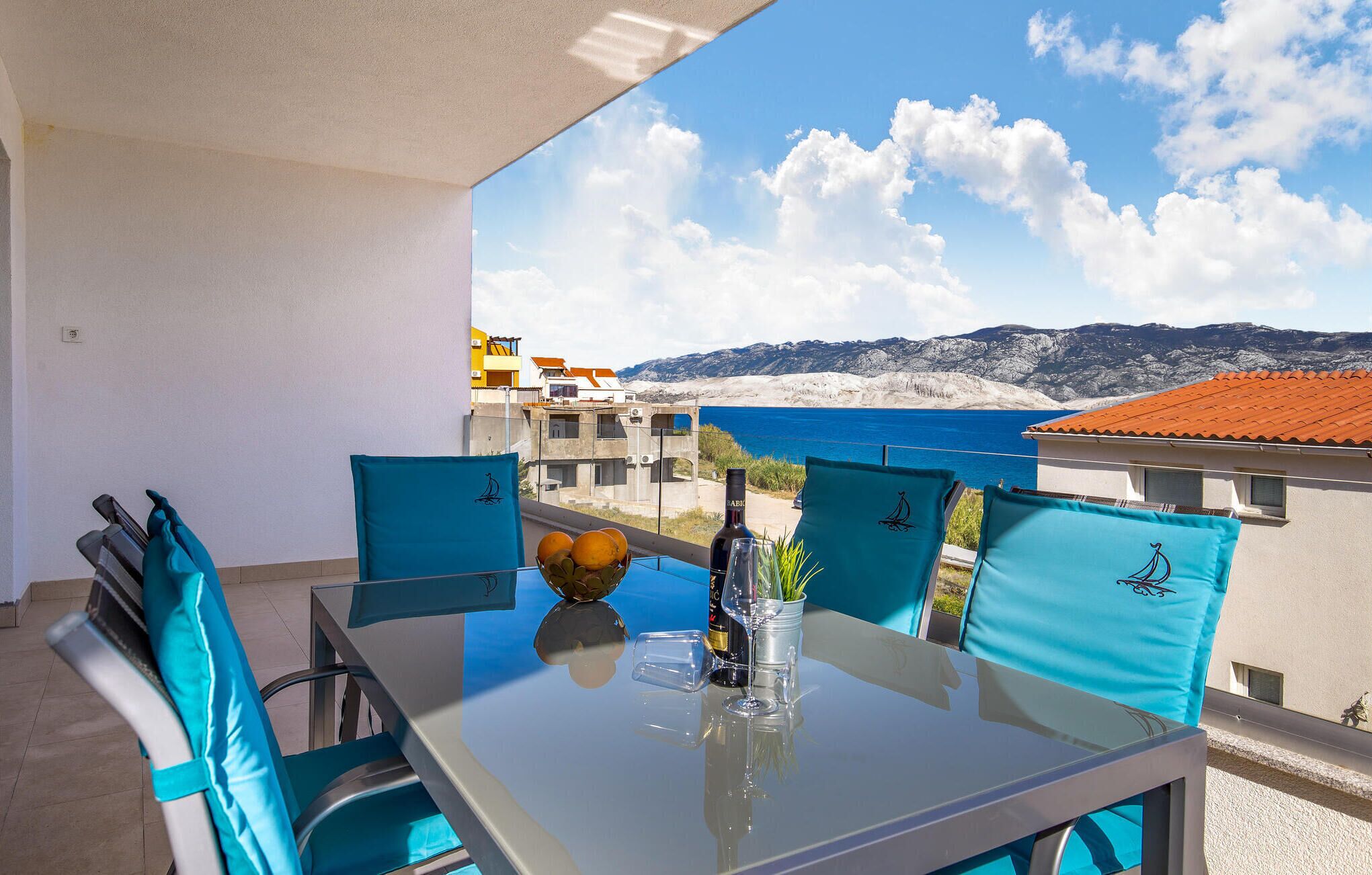 A balcony with a table set for two, featuring a bottle of wine, a glass, and a bowl of fruit. The view includes a coastal landscape with buildings and mountains.