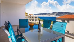 Un balcon avec une table dressée pour deux personnes, agrémentée d’une bouteille de vin, d’un verre et d’un saladier de fruits. La vue offre un paysage côtier avec des bâtiments et des montagnes.