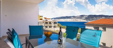 A balcony with a table set for two, featuring a bottle of wine, a glass, and a bowl of fruit. The view includes a coastal landscape with buildings and mountains.