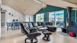 Een ruime woonkamer met een hoge nok, een grote zithoek, een eettafel en een open haard.