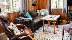 Un salon chaleureux avec un fauteuil en cuir, un canapé agrémenté de coussins à motifs, une table basse en bois et un téléviseur fixé au mur.