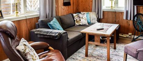 A cozy living room with a leather armchair, a sofa with patterned cushions, a wooden coffee table, and a TV on the wall.