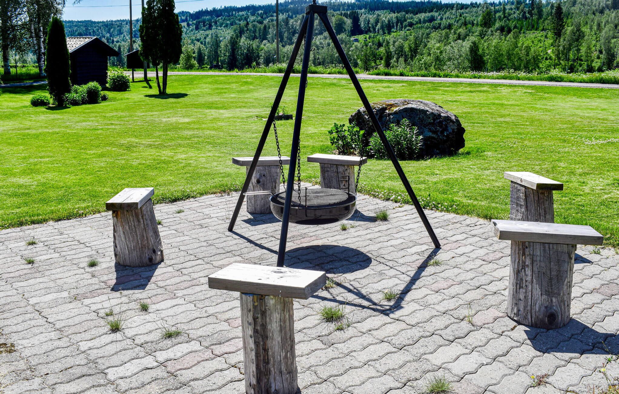 Une balançoire avec une structure en métal et des sièges en bois, entourée de piliers en pierre, installée sur une surface pavée, avec un champ d’herbe et des arbres en arrière-plan.