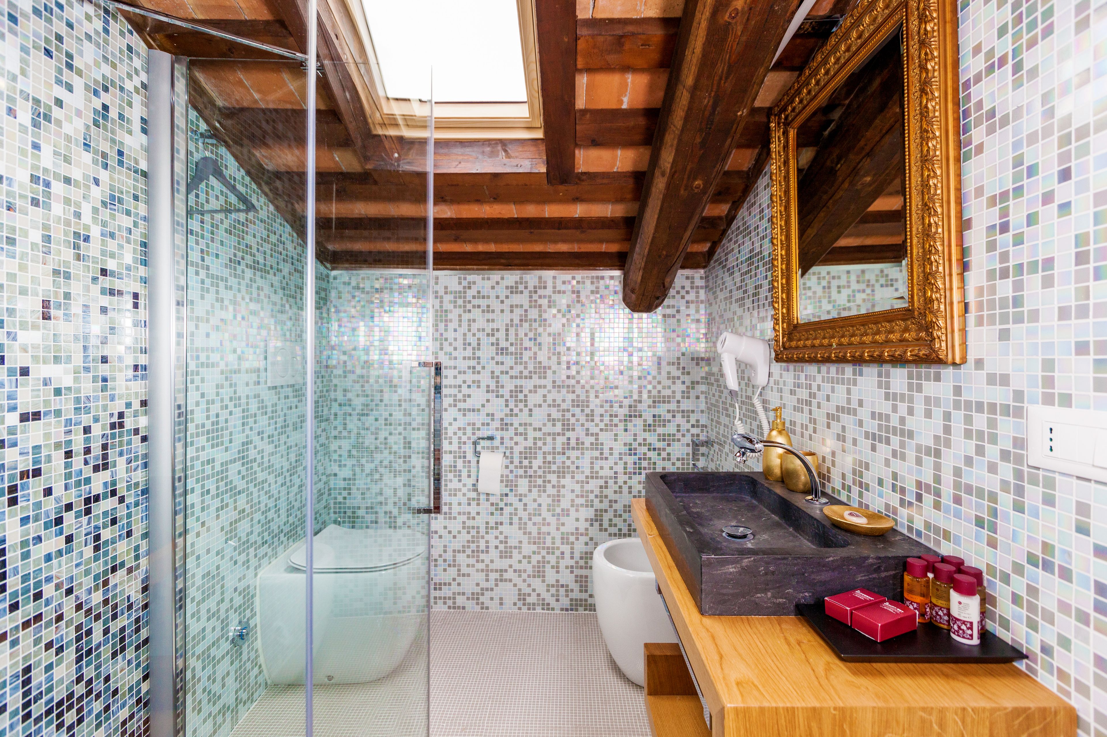 Casa de banho com paredes em mosaico, teto de madeira, cabine de duche em vidro, sanita branca, lavatório com bancada preta e espelho com moldura dourada.