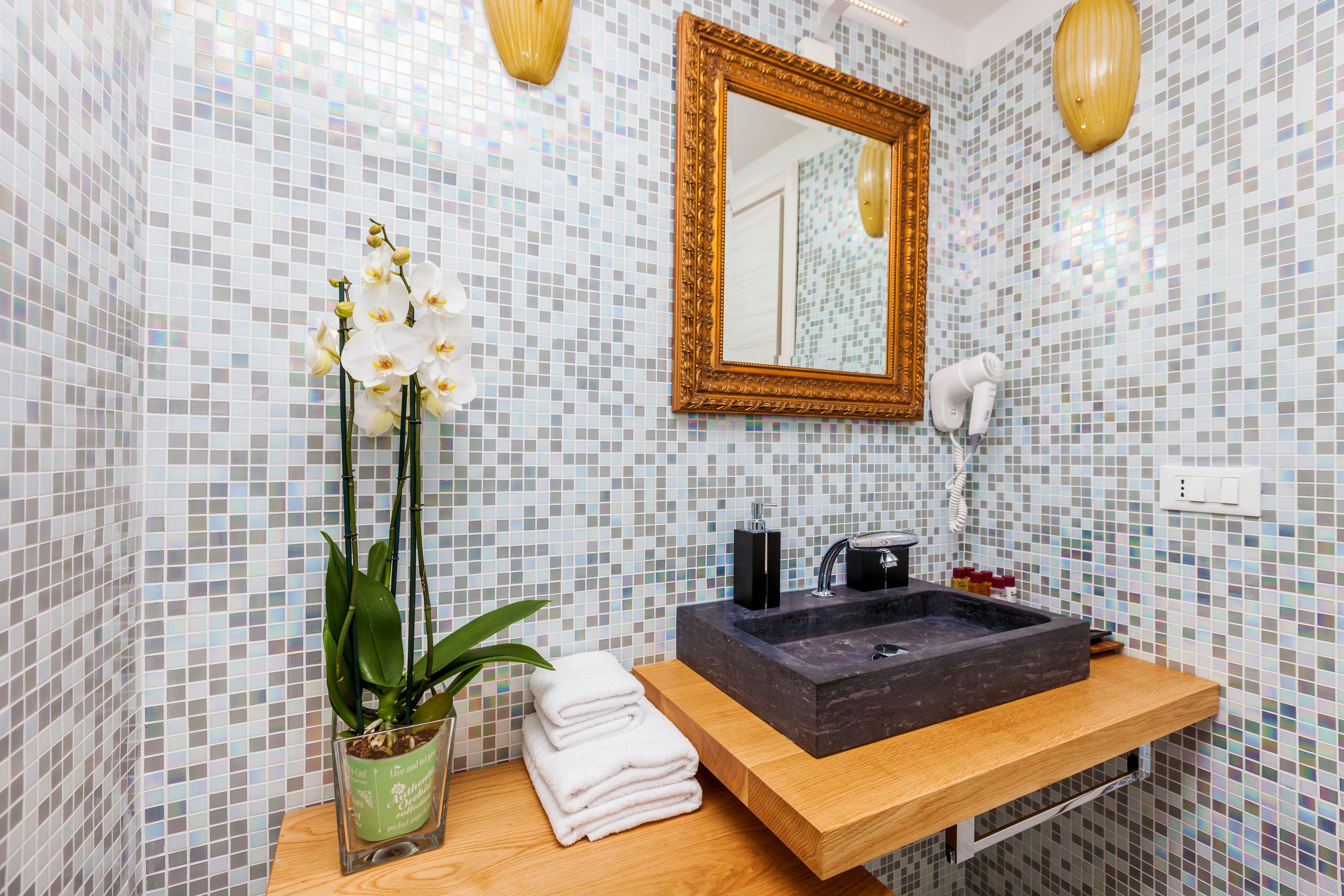 A bathroom with a mosaic tile wall, a wooden vanity with a black sink, a gold-framed mirror, and a potted orchid plant.