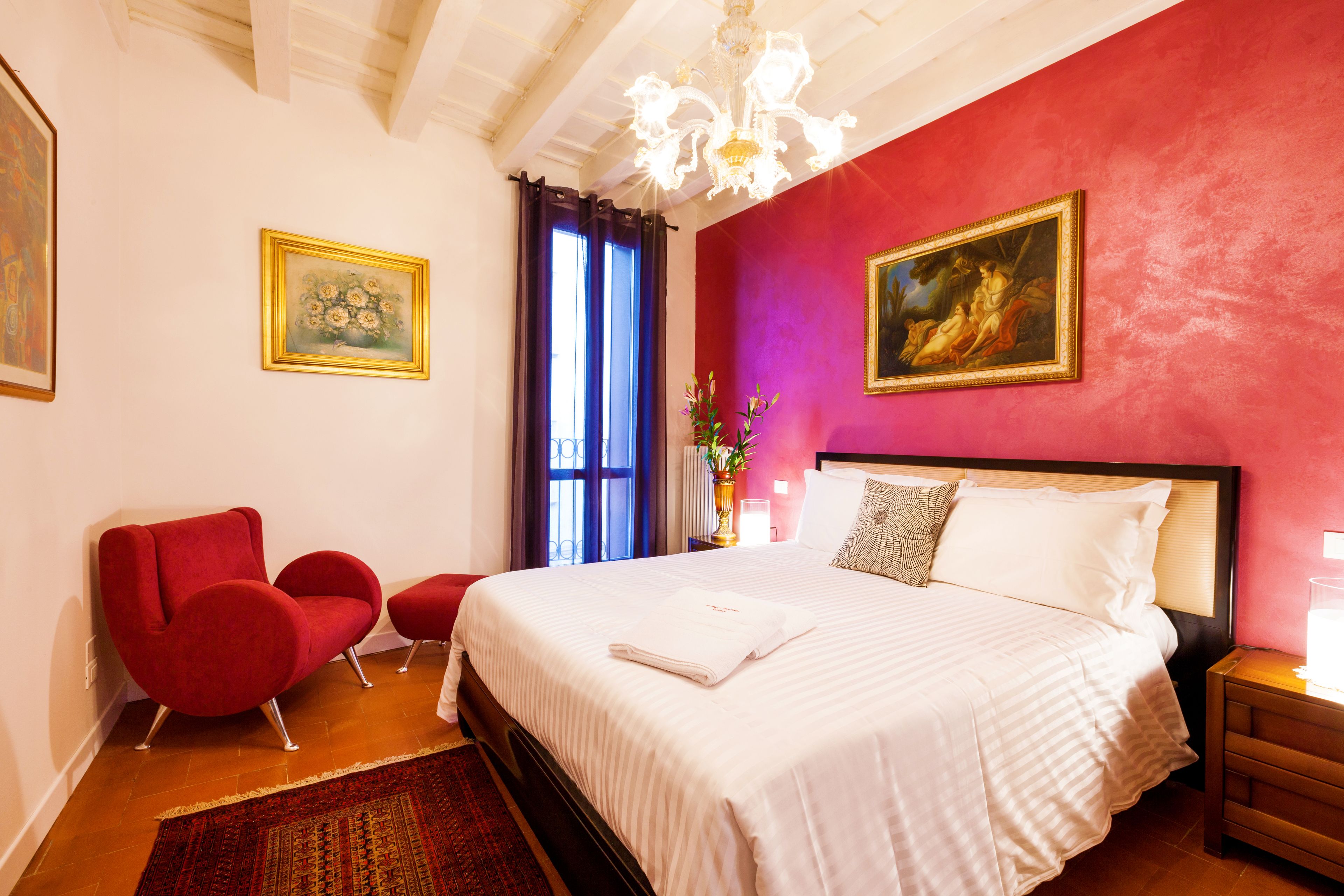 A hotel room with a large bed, red armchairs, a chandelier, and two paintings on the walls.