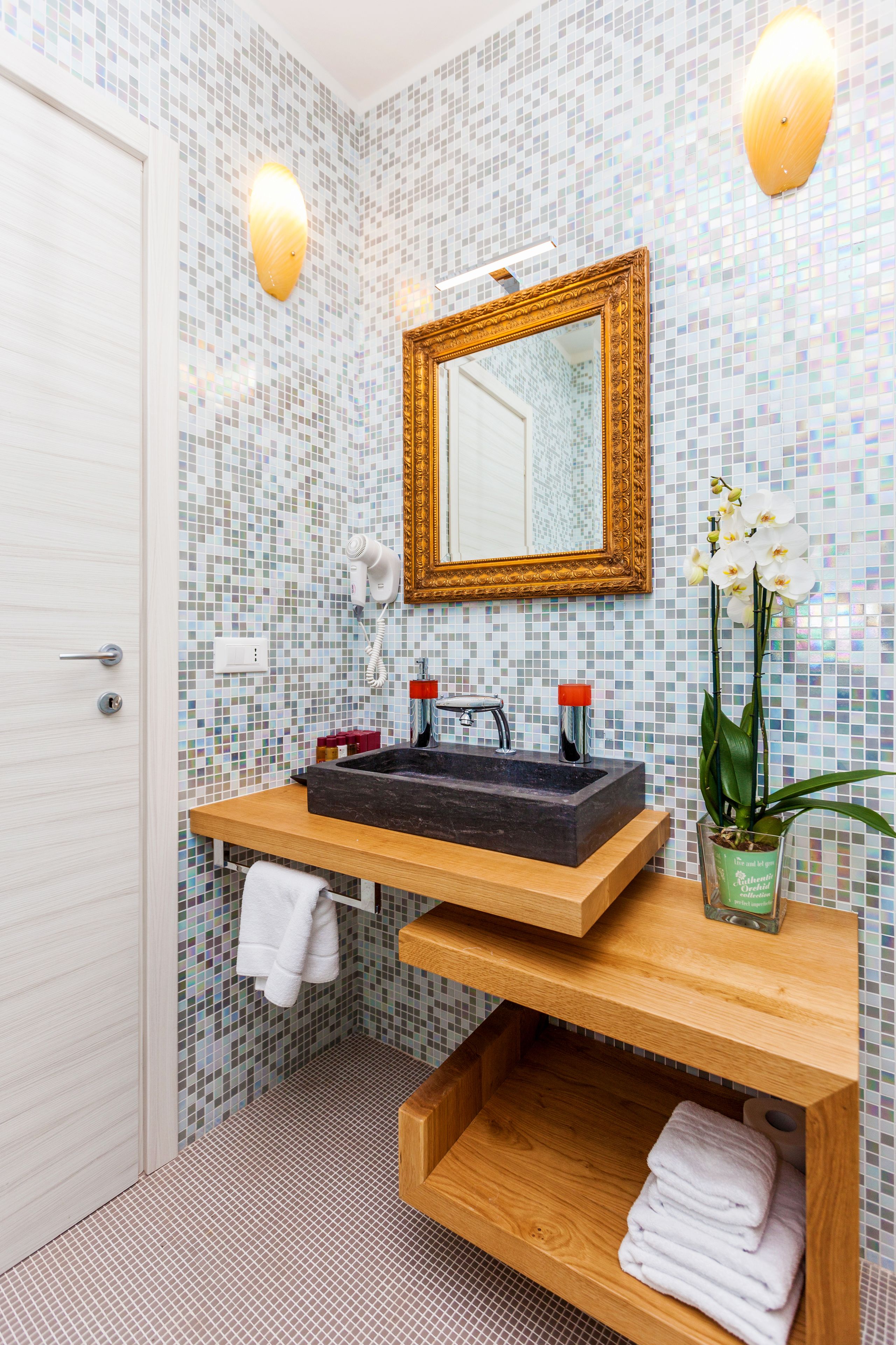 Un baño con pared de mosaicos, tocador de madera, lavamanos, espejo y una orquídea en maceta.