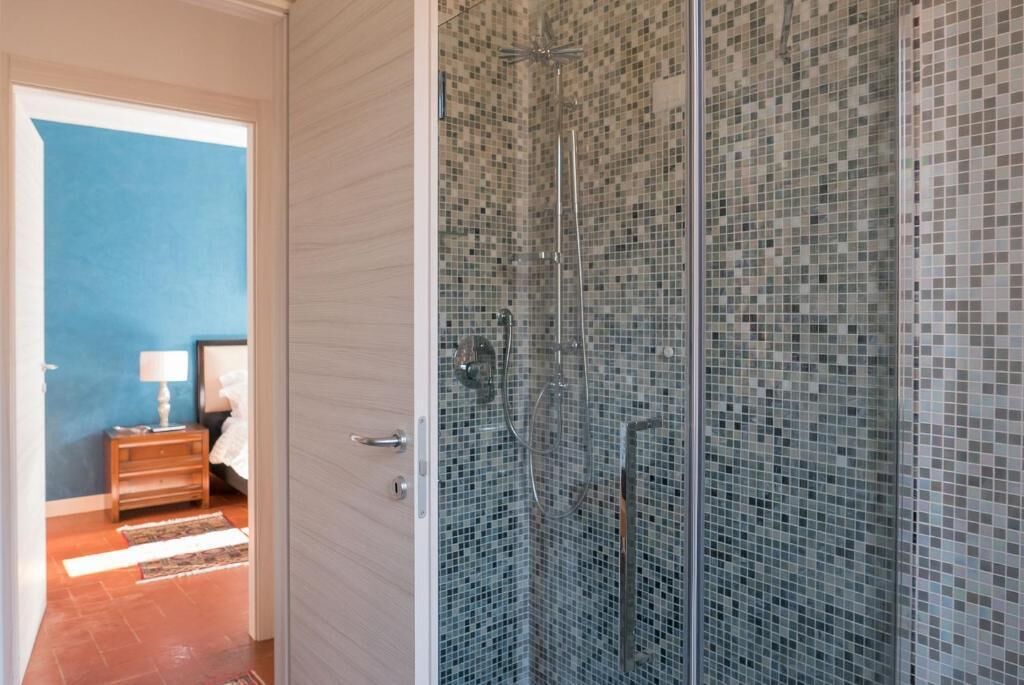 A glass-enclosed shower with mosaic tiles, a wooden nightstand with a lamp, and a blue wall in the background.