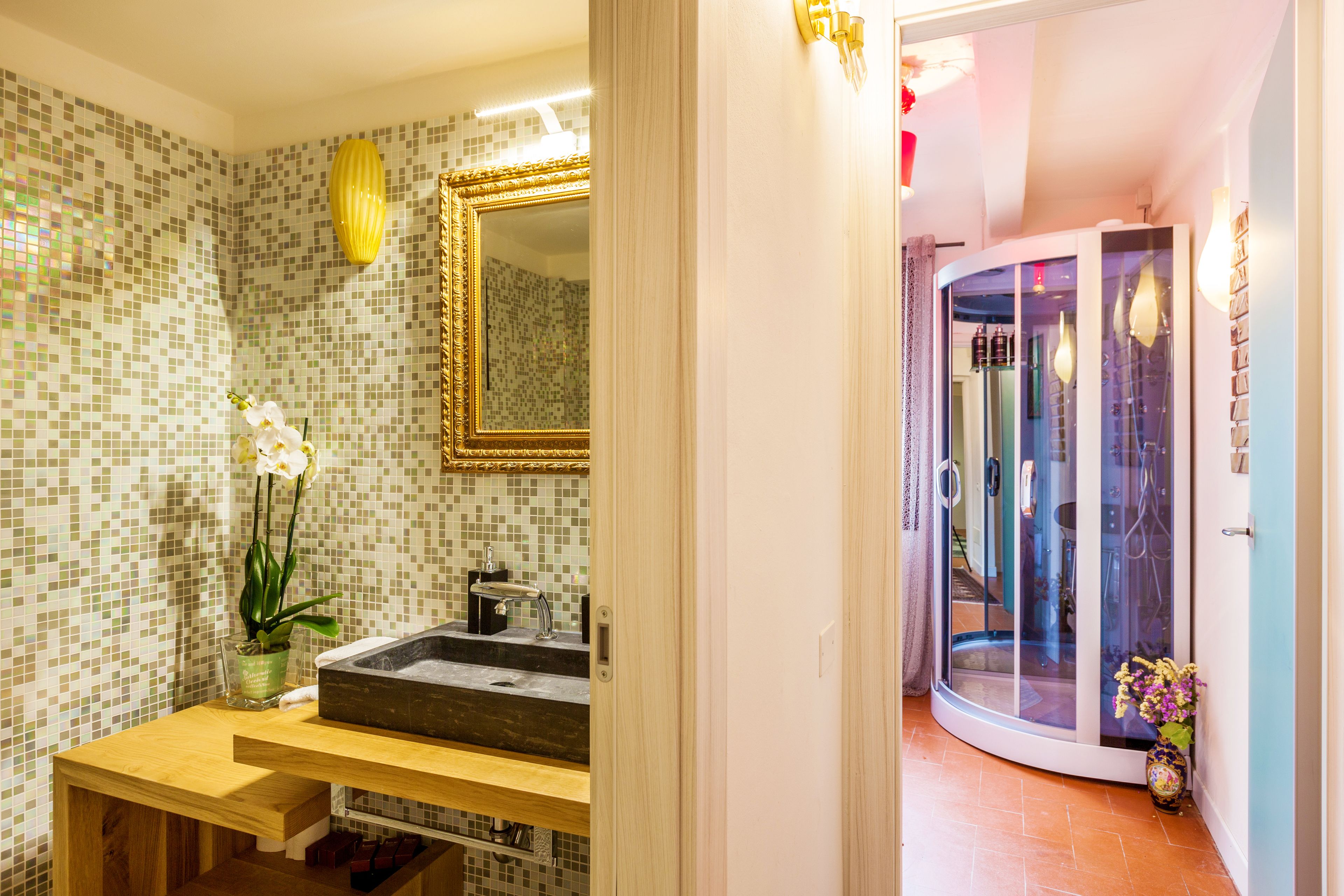 Ein Badezimmer mit Wand aus Mosaikfliesen, einem Holzkosmetikschrank, einem spiegel mit goldener Rahmung und einer Duschkabine aus Glas.