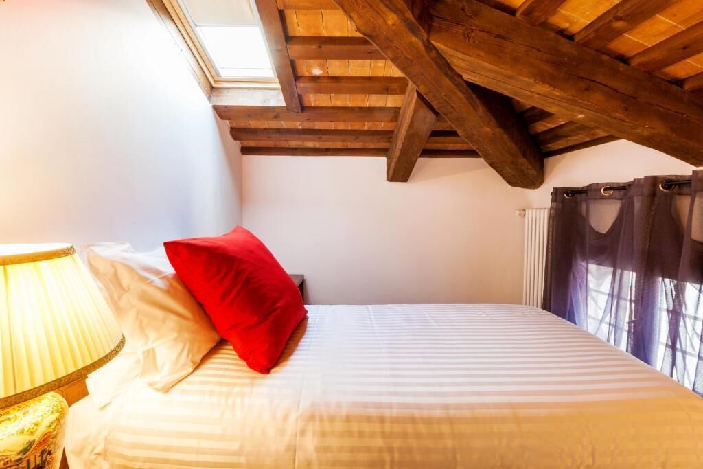 A bedroom with a wooden ceiling, a bed with white and red bedding, a lamp, and a window with curtains.