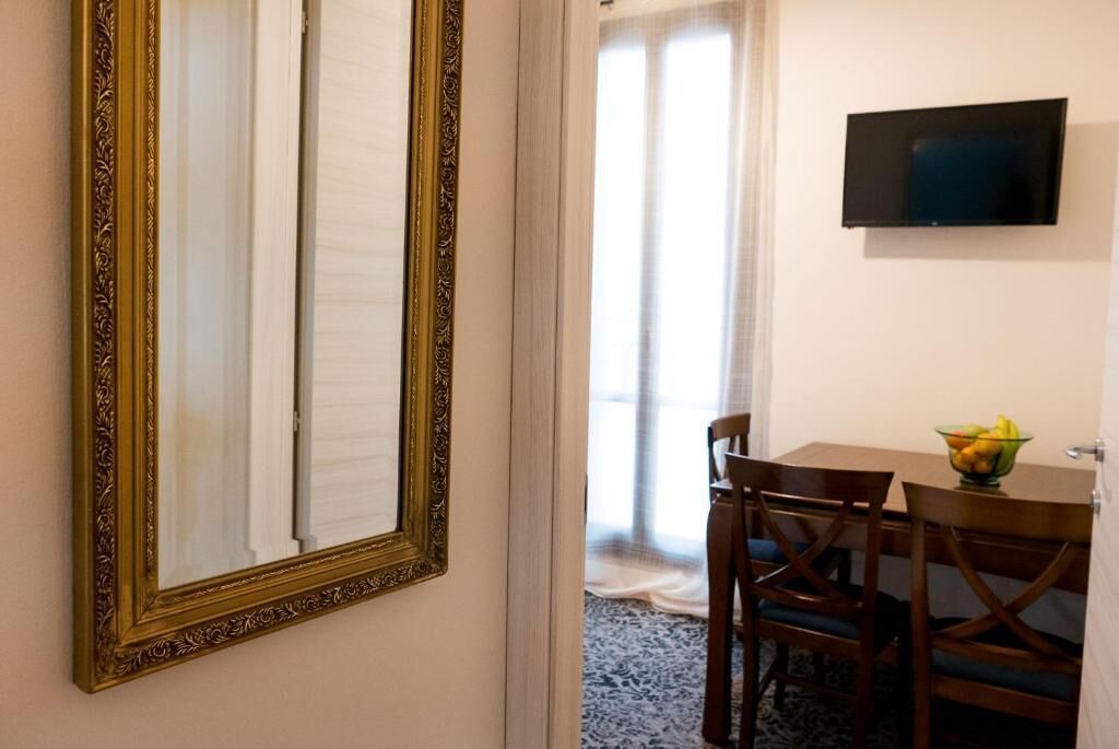 A room with a large ornate mirror, a flat-screen TV, a dining table with chairs, and a bowl of fruit.