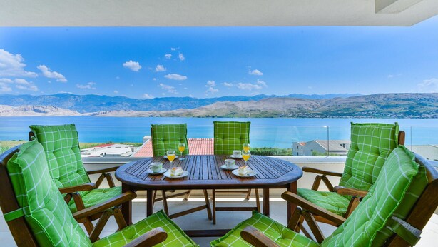 A round wooden table with four chairs, each with green cushions, set against a scenic backdrop of a lake and mountains. - Nice apartment in Pag with house sea view (Pag)