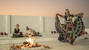 A person performing a dance with a colorful outfit, a fire on the ground, and two people sitting on steps in the background. - Dynasty Desert Camp (Jaisalmer)