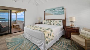 Un dormitorio con una cama grande, vista al océano, cabecera de madera, dos mesitas de noche con lámparas y una silla.
