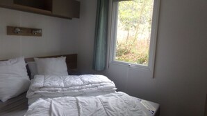 A bedroom with a bed, a window with a curtain, a shelf above the bed, and a wall-mounted light fixture.