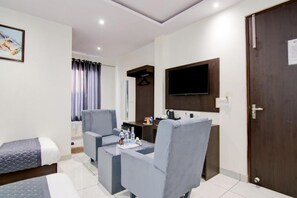 A hotel room with a bed, two armchairs, a small table with water bottles, a TV mounted on the wall, and a door with a sign. - Hotel De Hocks Deluxe (New Delhi)