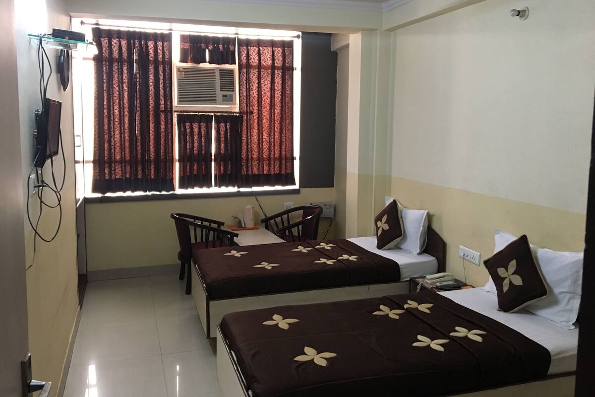 Habitación de hotel con dos camas, un escritorio, una silla, un ventanal con cortinas, aire acondicionado y ventilador de techo.