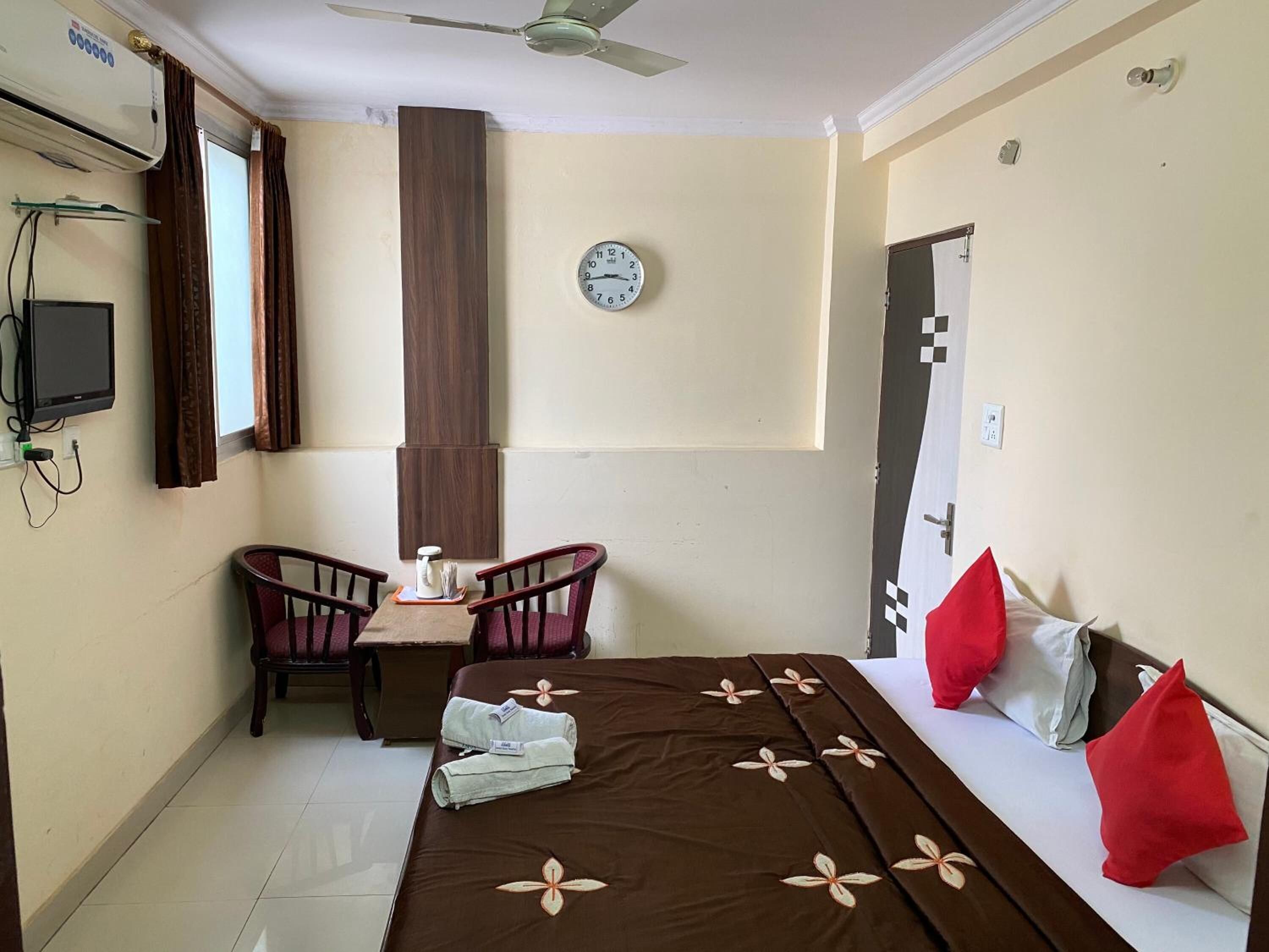 Habitación de hotel con una cama, un escritorio con dos sillas, un reloj en la pared y un ventilador de techo.