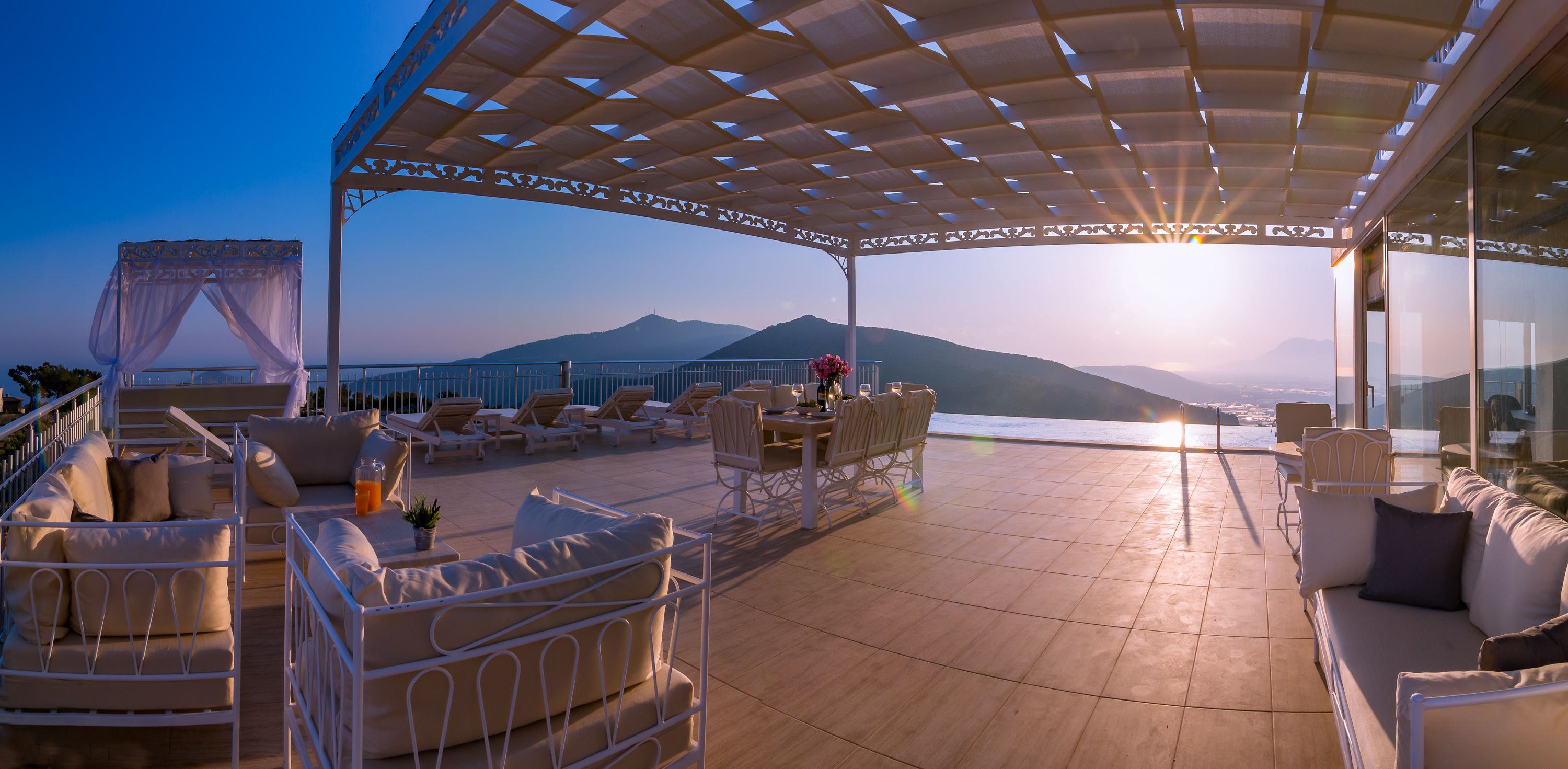 A sunlit outdoor lounge area with a view of mountains and the sea, furnished with white sofas and chairs, and a covered seating area.