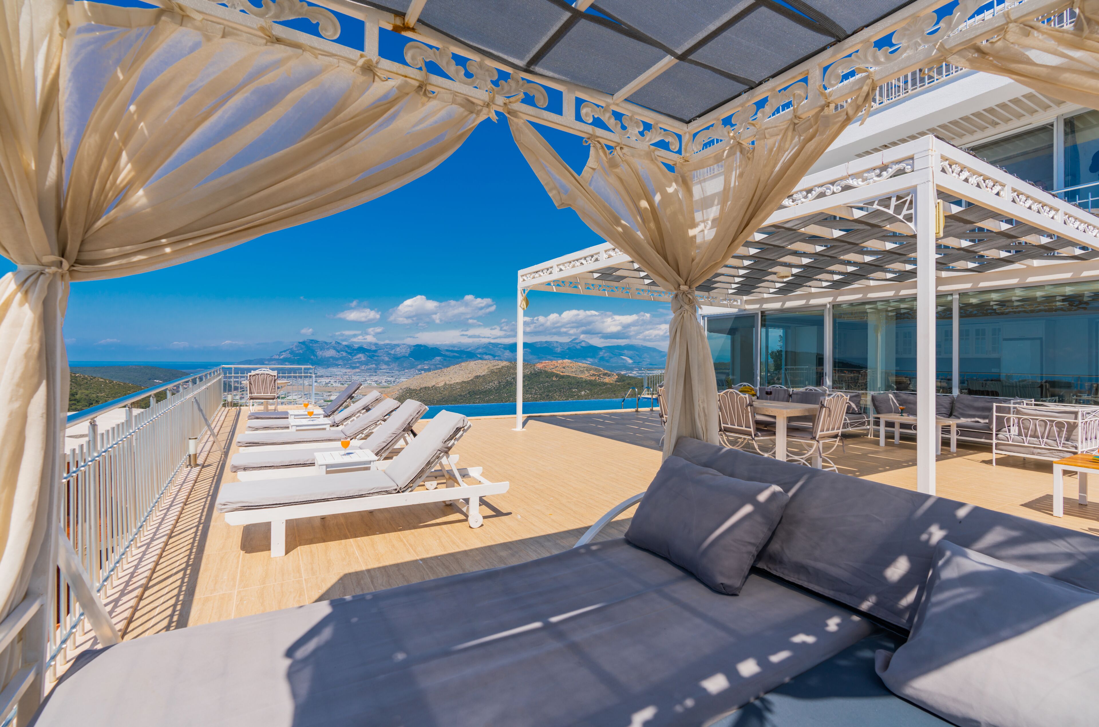 A sunlit outdoor lounge area with white lounge chairs and a sofa, a glass-enclosed dining area, and a view of the sea and mountains.