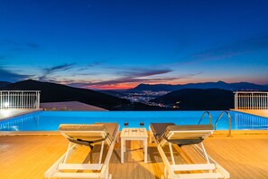 A poolside area with lounge chairs and a table, overlooking a cityscape and mountains at dusk. - Infinity Villa Kalkan (Kas)