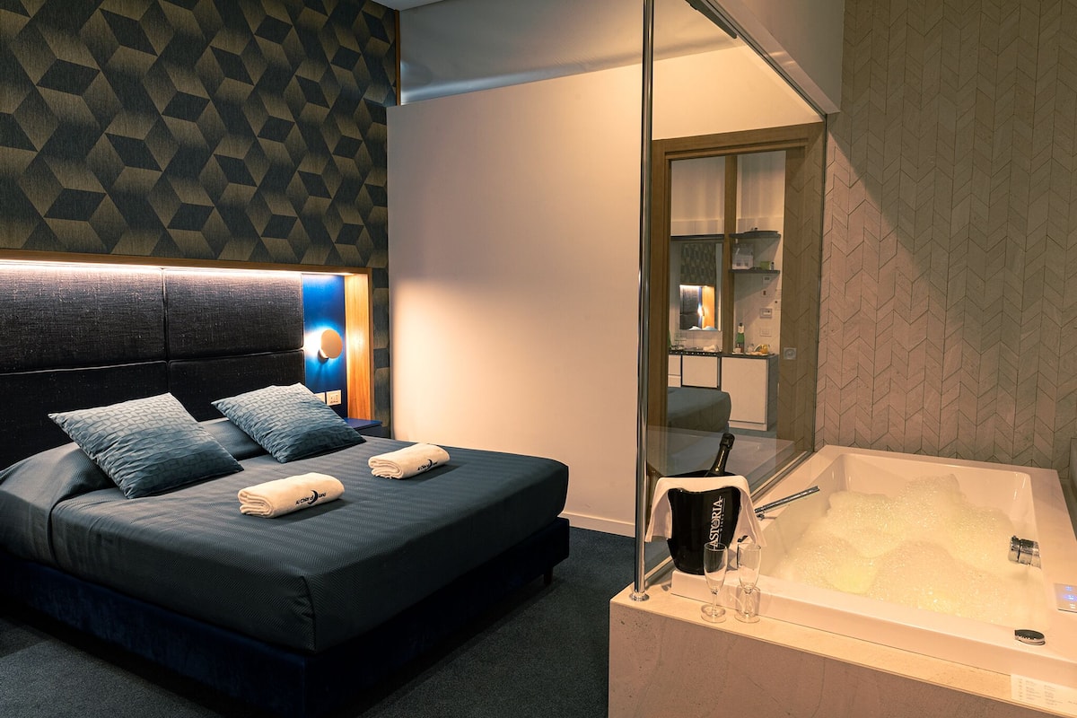 A modern hotel room with a large bed, a glass-enclosed bathtub, and a geometric-patterned wall.