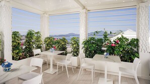 A modern outdoor dining area with white furniture and plants, overlooking a scenic view. - Al Chiar di Luna (Monte di Procida)