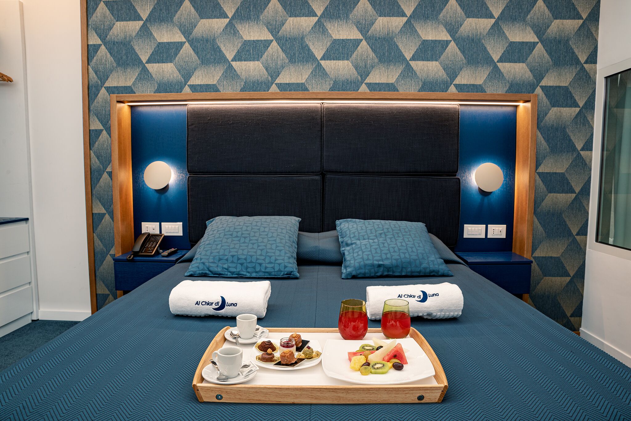 Ein Hotelzimmer mit einem großen Bett, zwei Nachttischen, einem Tablett mit Frühstücksgebäck und einer Wand mit Muster.