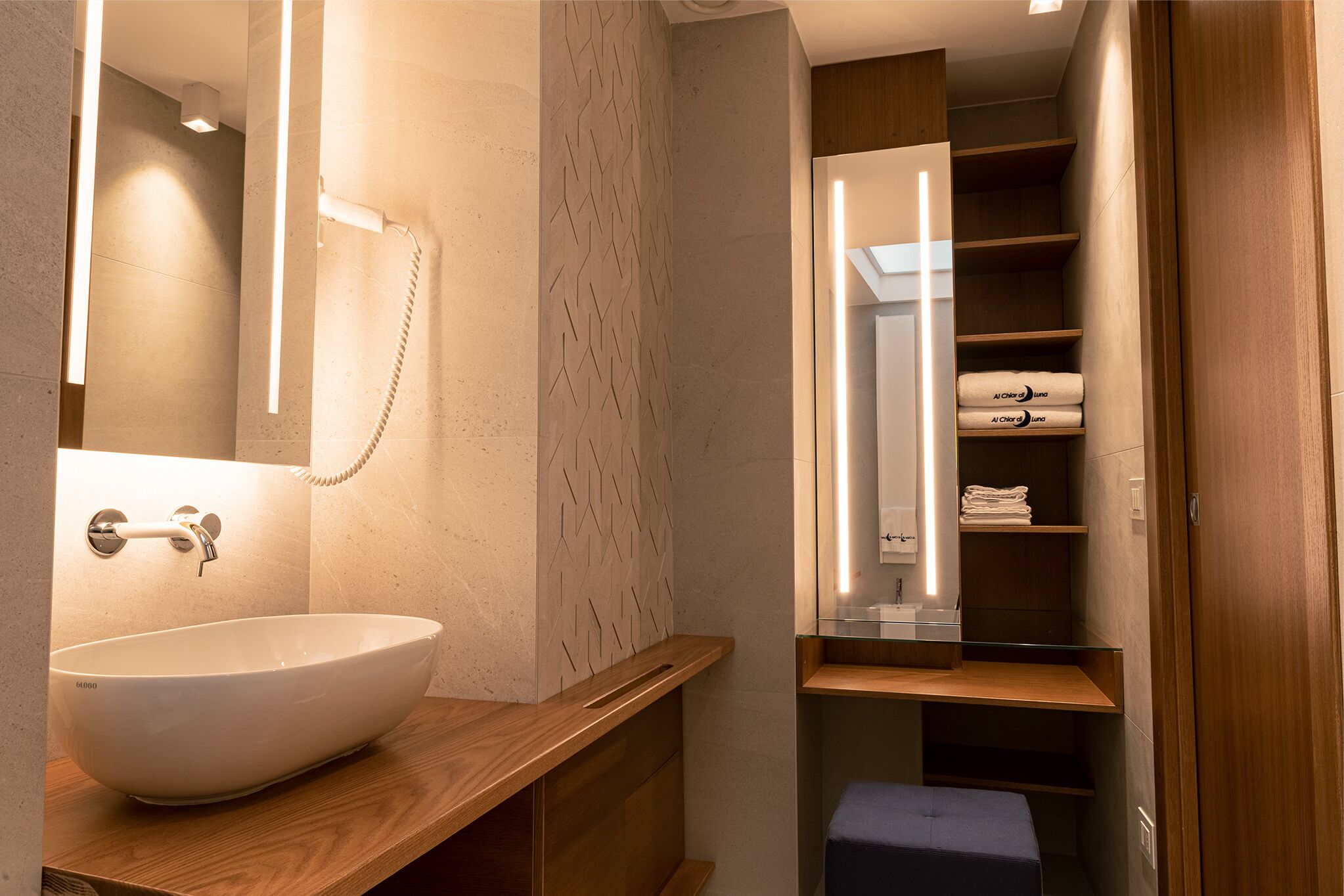 Un baño moderno con un lavamanos blanco tipo recipiente, un espejo y estantes empotrados con toallas dobladas.