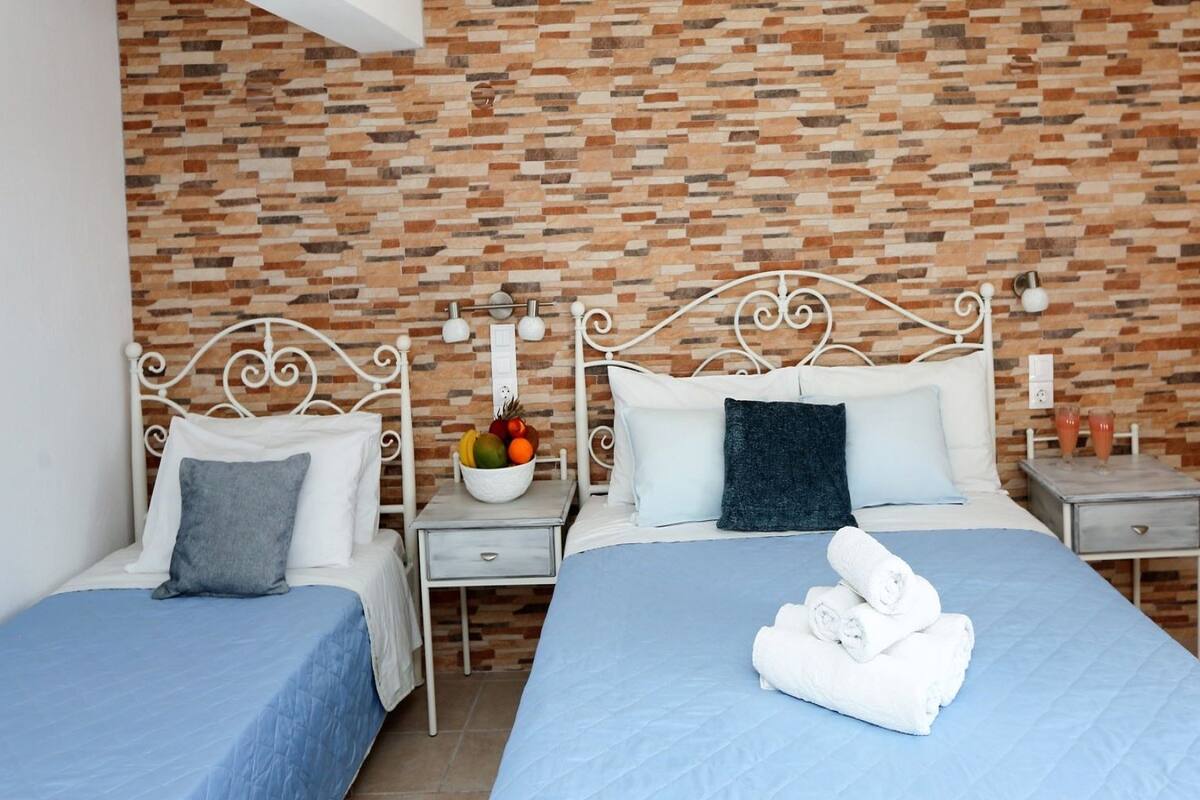A hotel room with two beds, a brick-patterned wall, white metal headboards, and a fruit bowl on the nightstand.
