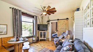 A cozy living room with a wooden floor, a sofa with patterned cushions, a wooden table with a bench, a ceiling fan, and a TV on a cabinet. - Walk to Dtwn Shops & Eats: Sturgeon Bay Retreat (Sturgeon Bay)
